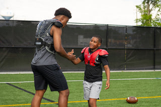 Two football players, one older and one younger, wearing Legion shoulder pads and shaking hands on a field