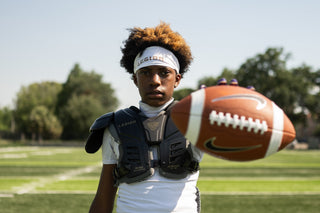 Young football player wearing a white LEGION Headband and shoulder pads, holding a football