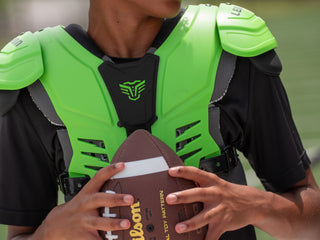 Person wearing green football shoulder pads holding a Wilson football on a field.