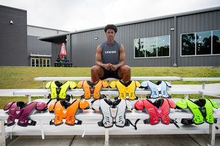 Young man sitting on bleachers surrounded by Legion M-80 All-Purpose Youth Shoulder Pads in various colors