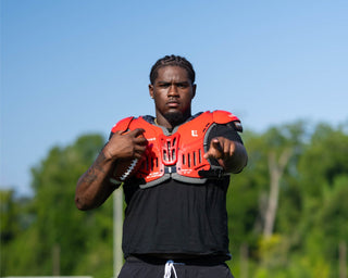 A man standing on a green football field wearing a black t-shirt, black shorts, and holding a red and black football shoulder pad in front of his chest.