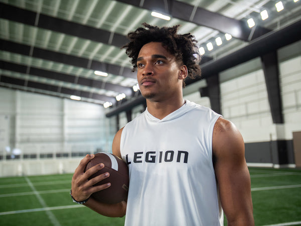 Athlete in a white Legion sleeveless hoodie holding a football on an indoor turf field
